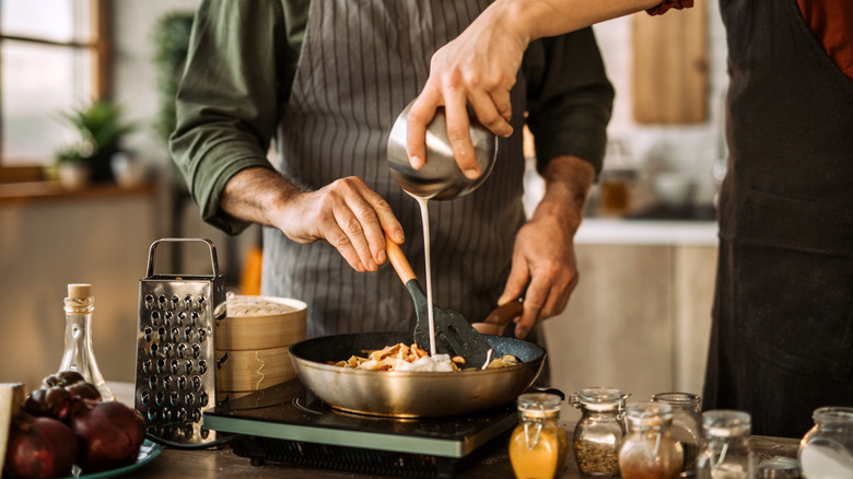 two cooks pour cream into pan of pasta on hot plate