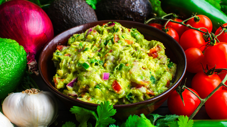 Bowl of guacamole surrounded by cherry tomatoes, avocados, lime, red onion, garlic and chilli peppers.