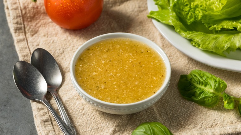A white bowl of salad dressing rests on a countertop near a plate of lettuce, spoons, and tomatoes