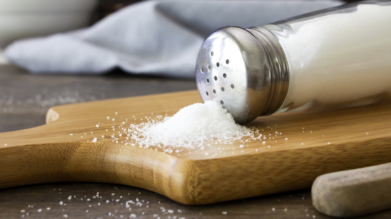 A salt shaker with a pile of salt beside it on a wooden kitchen board