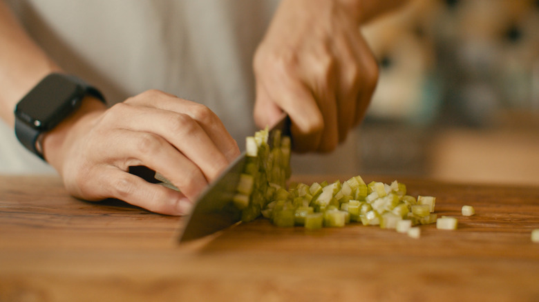 Hands dicing celery with a knife on a wooden surface