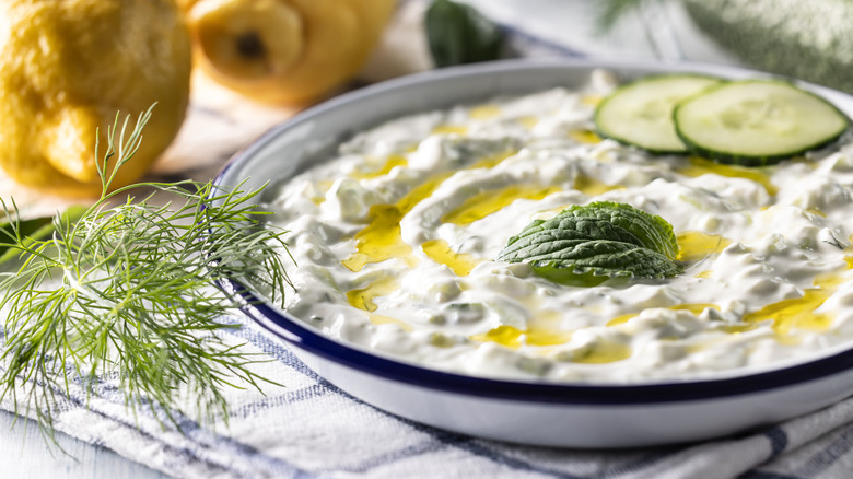 A bowl of tzatziki dip with lemons in the background, garnished with cucumber slices, mint, and dill