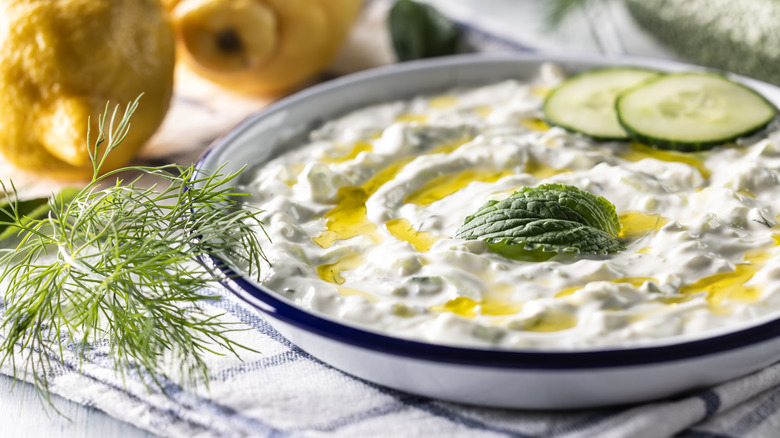 A bowl of tzatziki dip with lemons in the background, garnished with cucumber slices, mint, and dill
