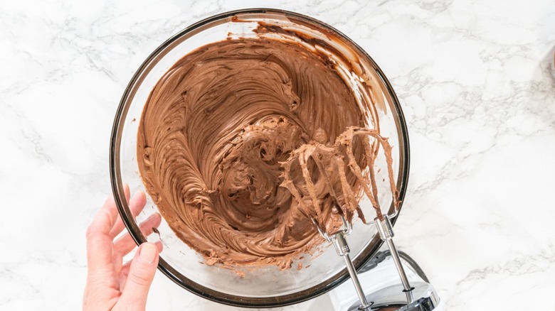 A person mixing chocolate frosting in a glass bowl.