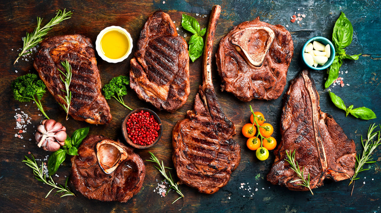 Grilled steak cuts on a wood table with fresh herbs and vegetables