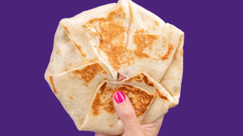 Hand holding a Taco Bell Crunchwrap Supreme in front of purple background