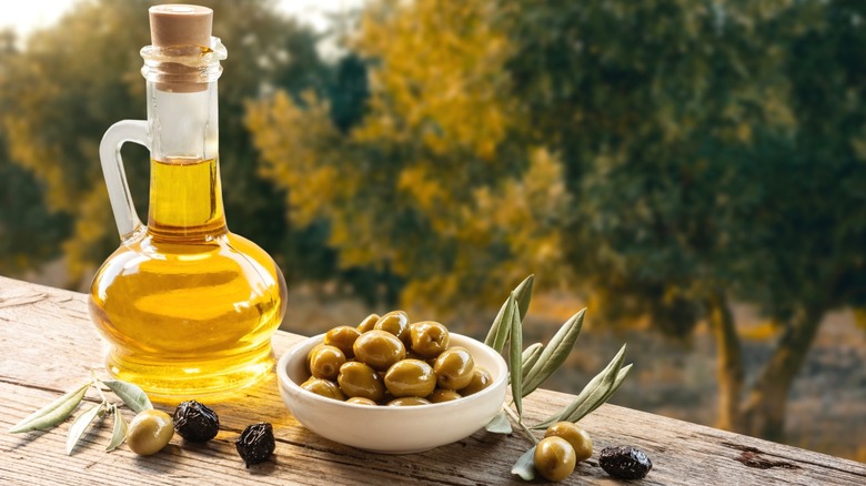 A jug of olive oil next to a bowl of olives in a garden.