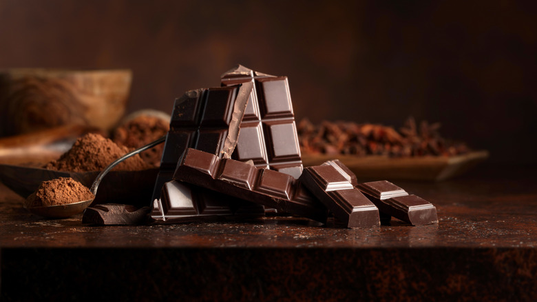 Cut up bars of chocolate on a wooden table with cocoa powder behind them.