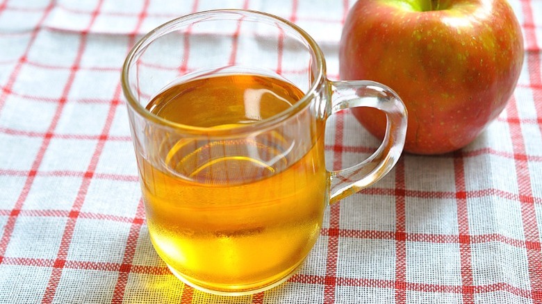 A glass cup of apple juice next to an apple on a checkered tablecloth