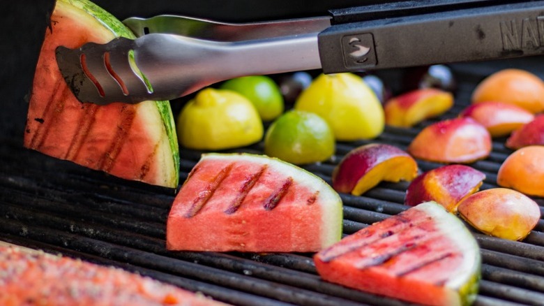 Tongs flipping slices of watermelon on a grill next to citrus fruit and peaches