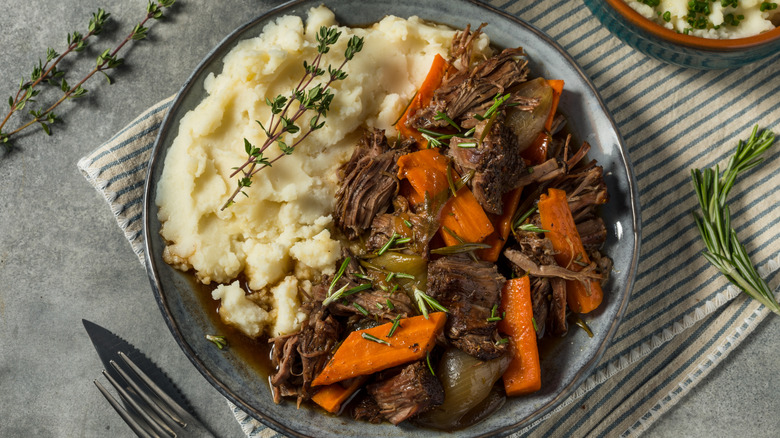 Plate of pot roast with carrots and onions alongside mashed potatoes with sprigs of thyme and rosemary