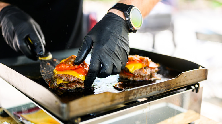 A pair of hands works with a spatula to make smash burgers on a flat griddle