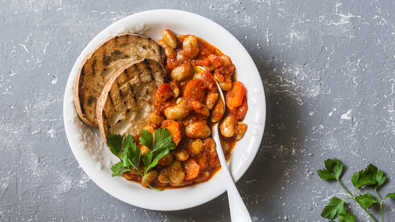 White bowl filed with gigande Italian beans, canned tomatoes and two pieces of grilled sliced bread on the side