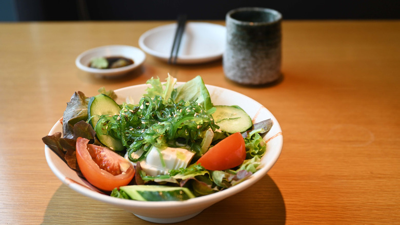 A fresh salad with tomato, seaweed, cucumber, and mixed lettuce at a Japanese restaurant in front of a bowl of soy sauce and wasabi and a tea mug