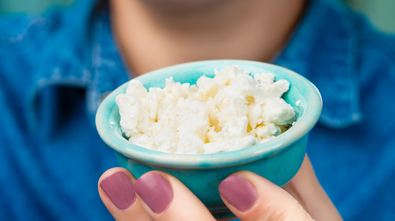 A person with mauve fingernails holding up a small blue bowl of cottage cheese