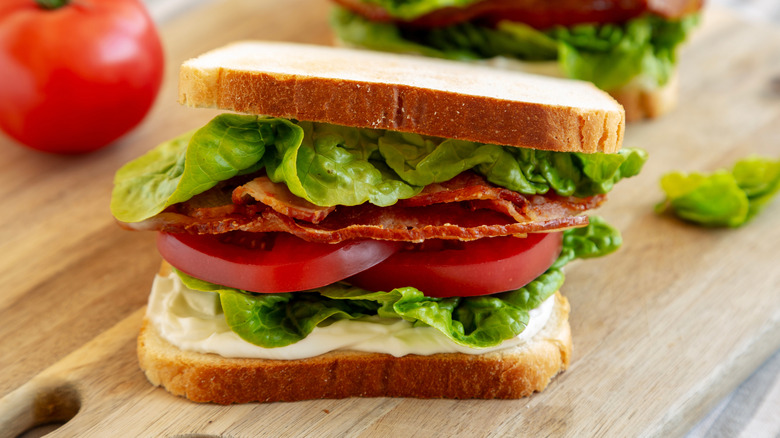 BLT sandwich with mayonnaise on a wooden surface near ingredients