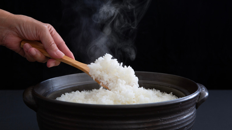 Someone spooning out some white rice from a clay pot on a black background.