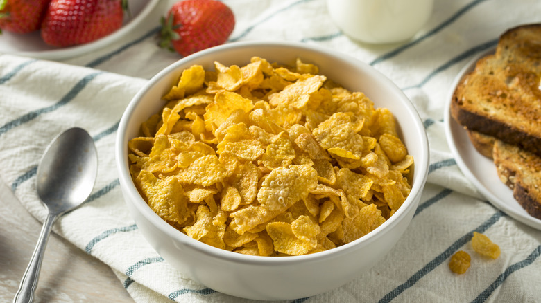 A bowl of cornflakes on a tablecloth next to strawberries, a spoon, milk, and toast