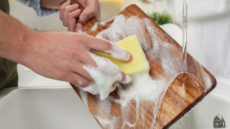 Hands washing a cutting board with soap and sponge over running water.