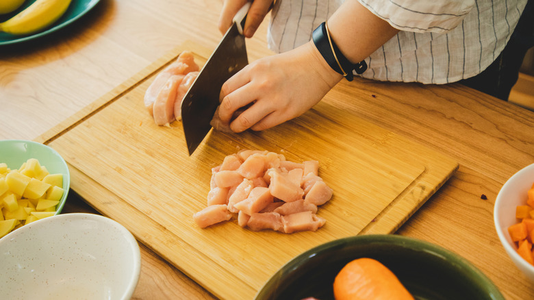 A person cutting chicken into cubes on a chopping board with a butcher knife as other plated ingredients are visible on their workstation.
