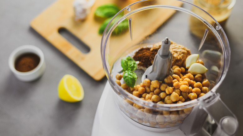 Overhead view of blender filled with hummus ingredients (chickpeas, basil, garlic) near a lemon half and a cutting board