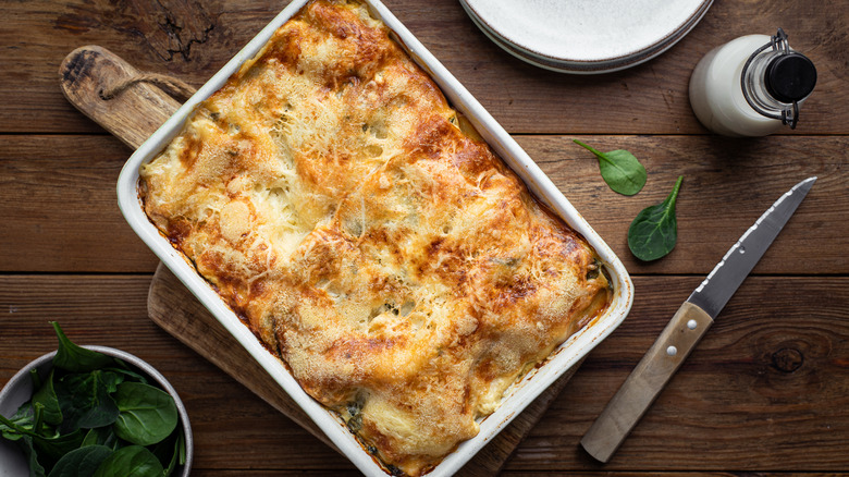 Lasagna in casserole dish on top of wooden serving board surrounded by knife, spinach, and plates