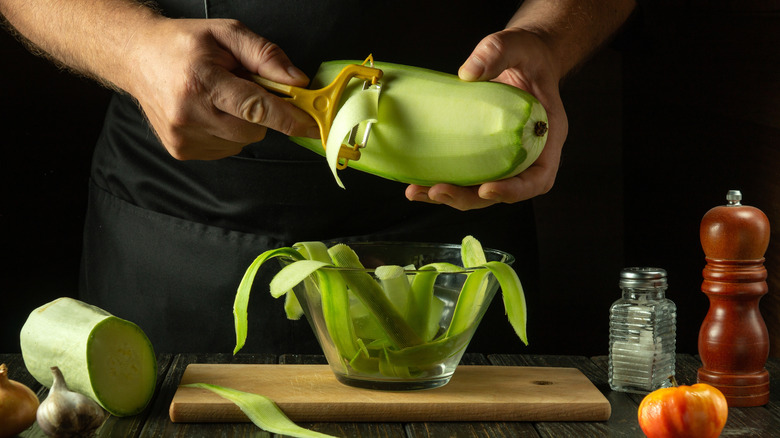 Hands peeling zucchini into thin strips into a glass bowl with a yellow vegetable peeler