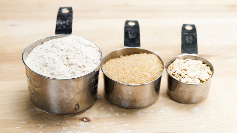 Three metal measuring cups of different sizes filled with flour, raw sugar, and oats