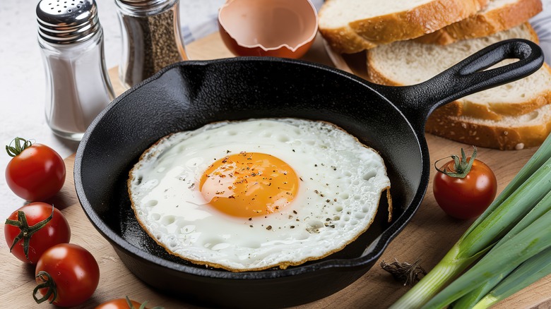 A single fried egg in a small cast iron skillet with tomatoes, toast, salt, and pepper