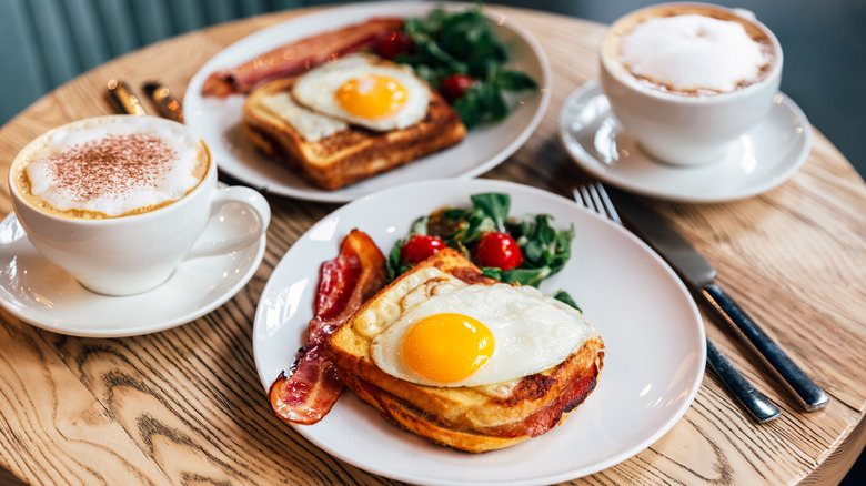 Two breakfast plates on a wooden table with fried eggs, toast, bacon, and side salad, with two lattes