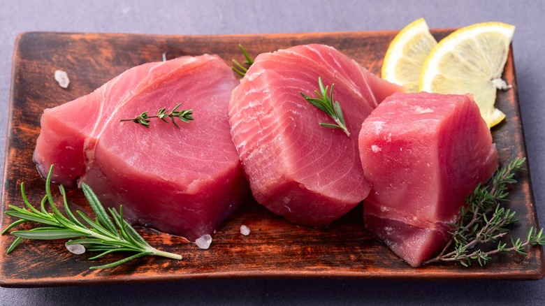 Three raw tuna steaks on wood board with herb sprigs and lemon slices