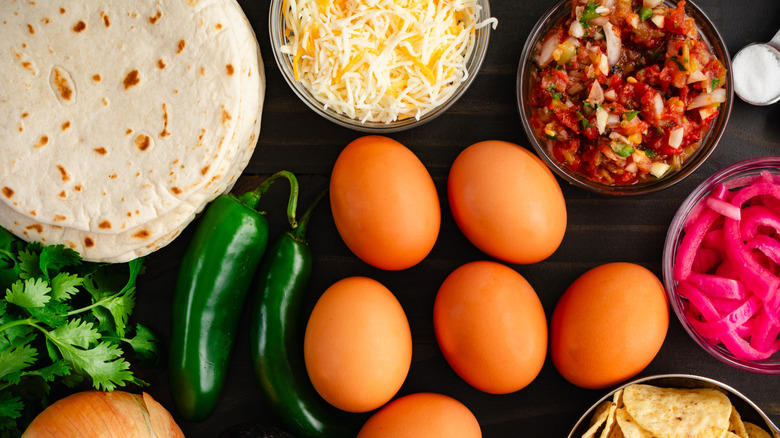 Top view of assorted Mexican breakfast ingredients including flour tortillas, shredded cheese, eggs, and salsa