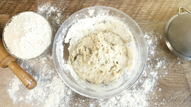 Glass bowl of dough with small glass bowl of flour nearby next to a rolling pin and a sifter on the opposite side