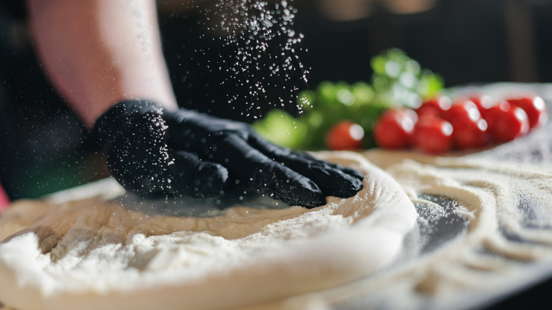 Hand with black glove on forming pizza dough with flour being dusted on top while tomatoes are in the background