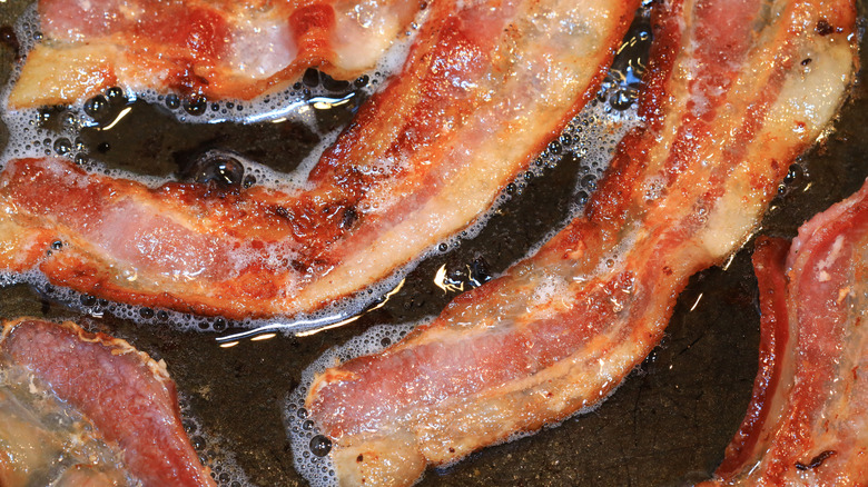 Close up of strips of bacon cooking in a pool of grease in a pot or pan.